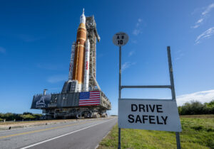 Space Launch System rocket on its way to the launch pad with "Drive Safely" sign in foreground