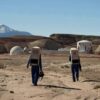 Two astronauts walking in the Utah desert with habitat buildings in the background