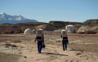 Two astronauts walking in the Utah desert with habitat buildings in the background
