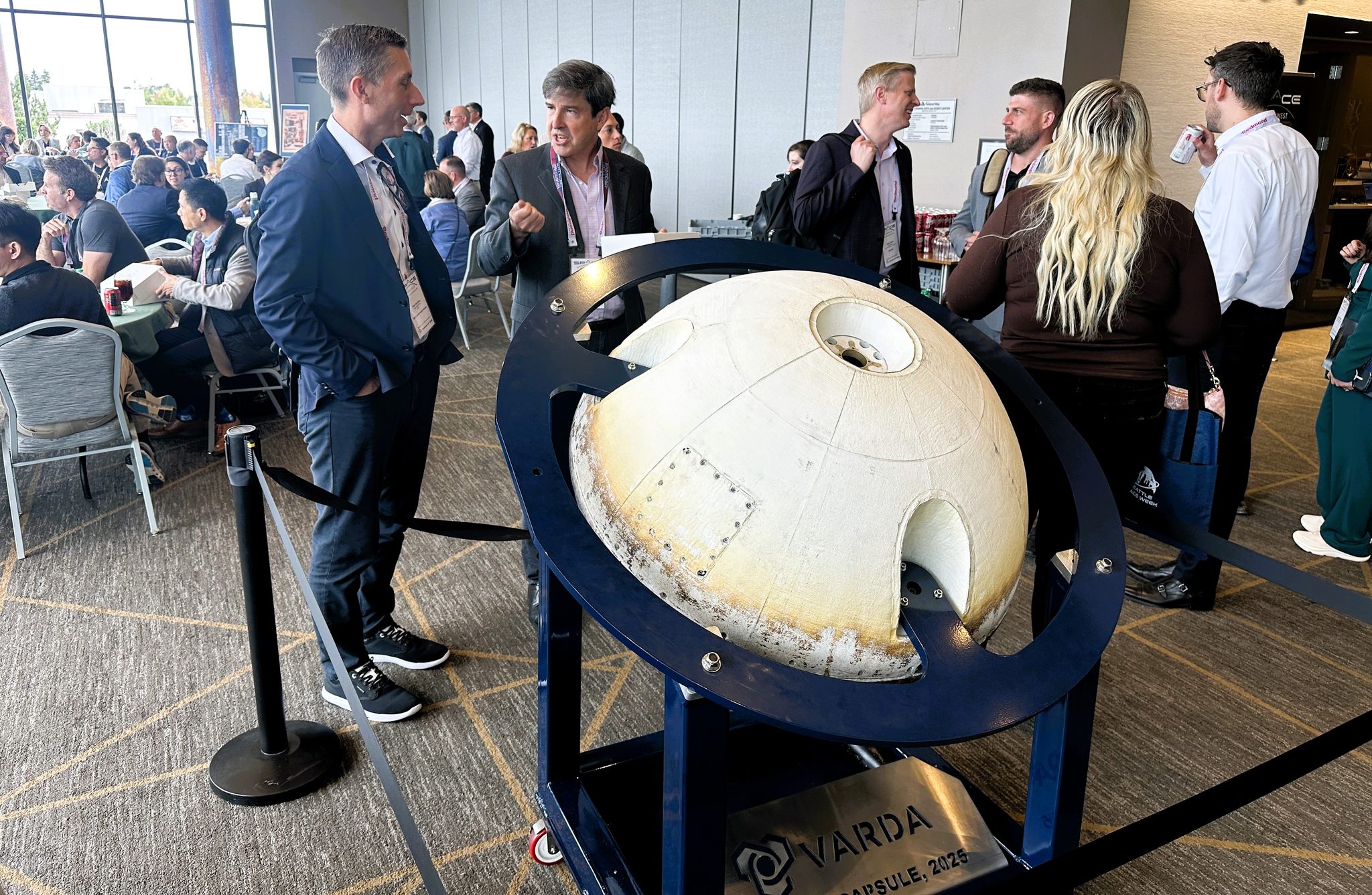 Dale Goulding, chief operating officer at Forge Digital, chats with Michael Reilly, chief strategy officer at Varda Space Industries, at the Space Northwest Symposium in Federal Way, Wash. One of Varda’s orbital capsules is on display in front of them. (GeekWire Photo / Alan Boyle)
