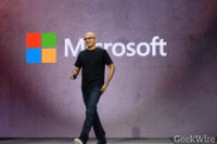 Satya Nadella walks across a stage in front of a large Microsoft logo.