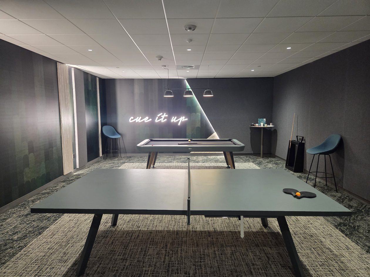 A dimly lit game room with a ping-pong table in the foreground, a pool table behind it and a lighted neon sign on the back wall.