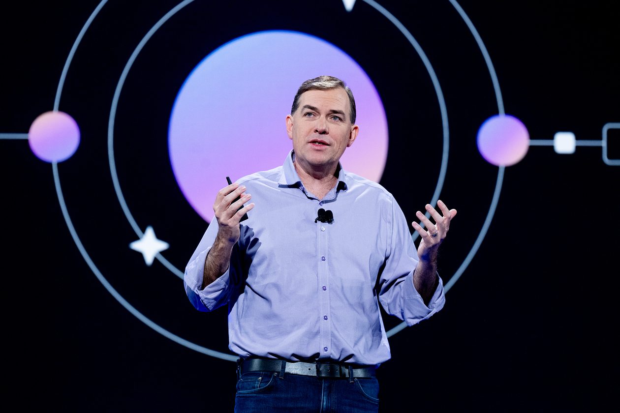 Matt Garman speaks on stage in a light purple button-up shirt, gesturing with both hands and holding a presentation clicker, in front of a dark screen showing a purple circle with orbit lines.
