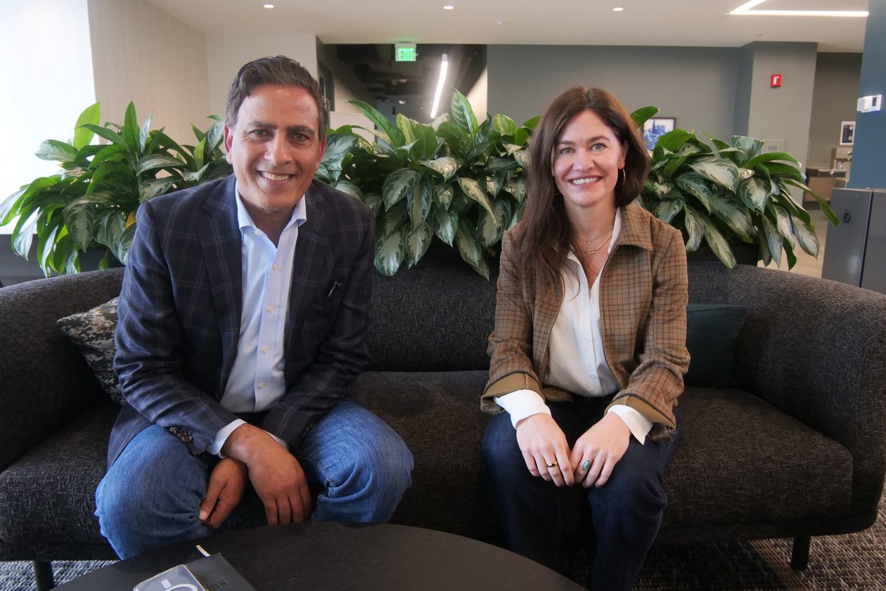 Ture Armas and Kristine Baker sit side by side on a dark gray couch in front of a row of leafy green plants in an office lounge.