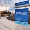 A blue Zillow Homes for-sale sign in front of a house on a residential street.