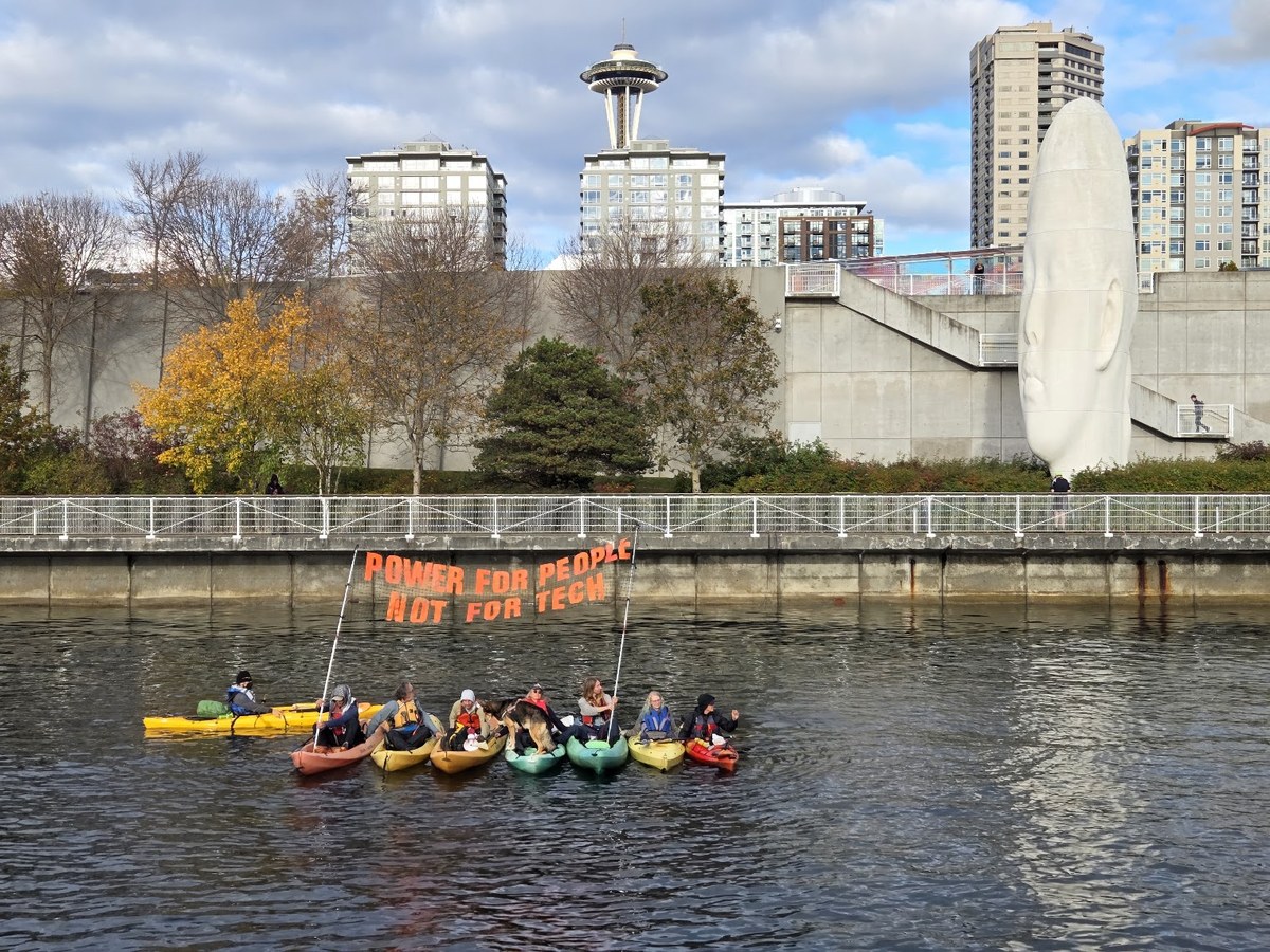 Protesters in kayaks on Elliott Bay hold a banner reading Power for People Not for Tech, with the Space Needle in the background.