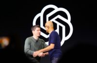 Sam Altman and Satya Nadella shake hands on stage in front of the OpenAI logo