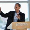 Andy Jassy gestures with both hands while speaking at a podium with the Amazon logo, in front of large windows.