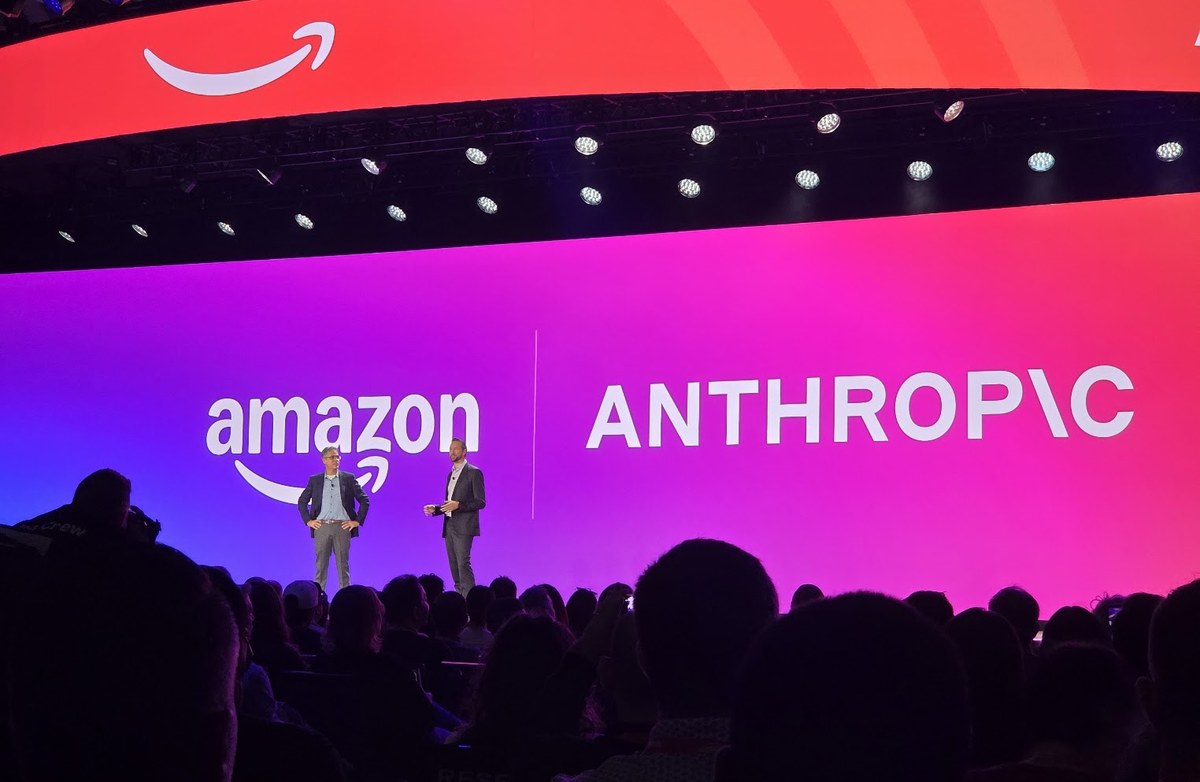 Two men stand on a stage in front of large screens showing the Amazon and Anthropic logos, with an audience in the foreground.