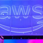 Illuminated AWS logo with the Amazon smile arrow on a glass wall in blue and purple light.