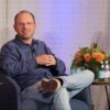 Thomas Dohmke, wearing glasses and a blue shirt, smiles while seated in an armchair on stage during a panel discussion.