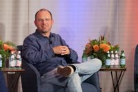 Thomas Dohmke, wearing glasses and a blue shirt, smiles while seated in an armchair on stage during a panel discussion.