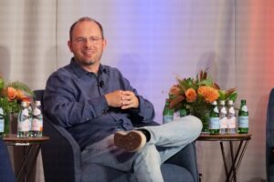 Thomas Dohmke, wearing glasses and a blue shirt, smiles while seated in an armchair on stage during a panel discussion.
