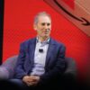 Amazon CEO Andy Jassy smiles while seated onstage in front of a red screen showing the Amazon logo.