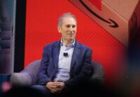 Amazon CEO Andy Jassy smiles while seated onstage in front of a red screen showing the Amazon logo.