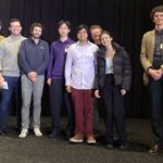 Eight people stand on a stage in front of a black curtain