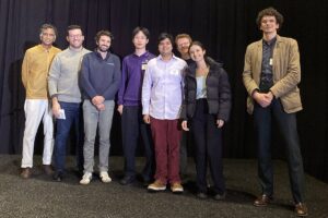 Eight people stand on a stage in front of a black curtain