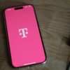 A smartphone showing the T-Mobile logo on a pink screen, lying on a wooden table.